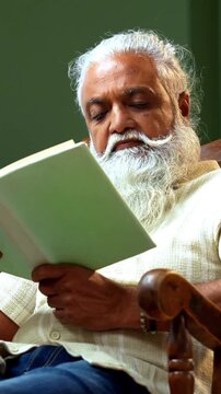 Indian mid age bearded man reading book novel on relaxing chair at home, stylish mature male enjoying peaceful leisure time with printed copy in modern interior lifestyle setting