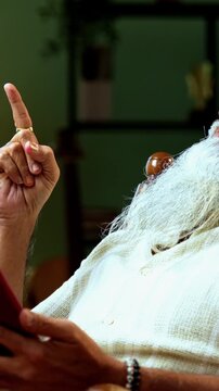 Indian mid age bearded man watching movie using smartphone sitting on relaxing chair at home, stylish mature male enjoying comedy short videos or reels with headphones in modern interior