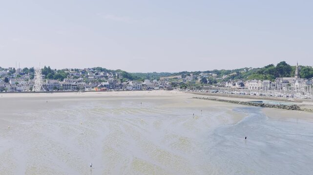 Aerial view of Binic beach and town on a clear summer day in Brittany, France with the expansive sandy beach at low tide, with shallow water patterns, a few people scattered across the sand.
