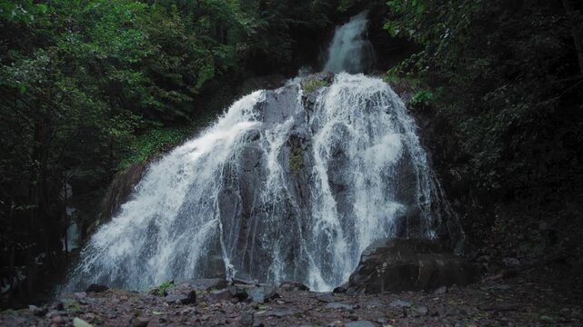 Dramatic cascading waterfall flowing over dark rocks in a lush green forest environment captured in a short video sequence