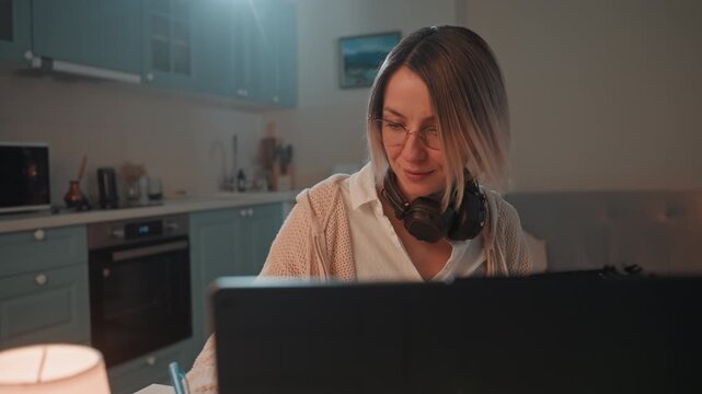 White woman music lover composing at laptop headphones around neck lamp casting warm glow cozy cardigan jotting lyrics in notebook nighttime kitchen studio focused smile intimate creative scene