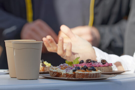 Takeaway coffee cups and gourmet tartines topped with cream cheese, olives and red onion sit on a table at Naplavka farmers market in Prague, while friends chat with lively hand gestures.