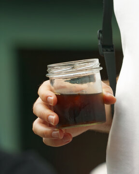 Fingers grip a repurposed glass jar filled with iced coffee, condensation beading on the rim, photographed in tight close-up at Naplavka farmers market in Prague.
