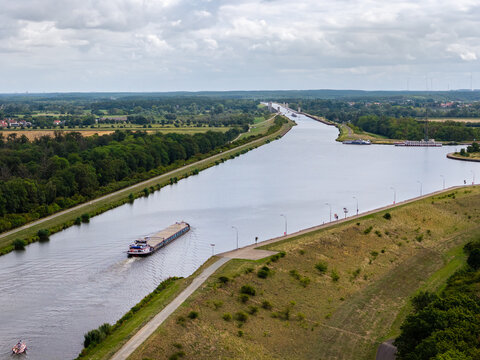 Cargo barge navigates broad Mittellandkanal near Magdeburg, surrounded by green countryside and canal infrastructure. Aerial view shows inland shipping, vessels, waterways and distant wind turbines