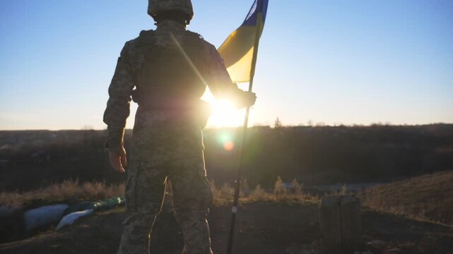 Rear view of male soldier in military uniform stands to the peak of hill with waving flag of Ukraine. Ukrainian army man lifting blue-yellow banner against sunset. Resistance to russian invasion