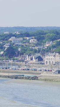Wide view of Binic beach and town in Brittany, France, showing a marina filled with sailboats, traditional coastal houses on a green hillside, and a sandy shoreline at low tide.
