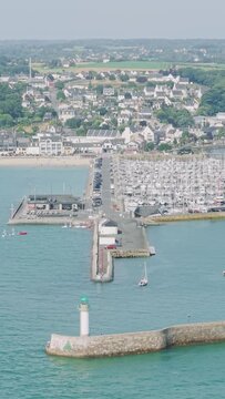 High-angle aerial footage of Binic, a coastal town, showing the marina filled with numerous boats, a stone jetty with a green-topped lighthouse in Brittany, France.