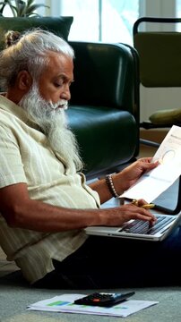Indian mid age bearded man using laptop, calculator and reports for accounting sitting on carpet in modern home, stylish mature male managing finance, budget and financial statements indoors