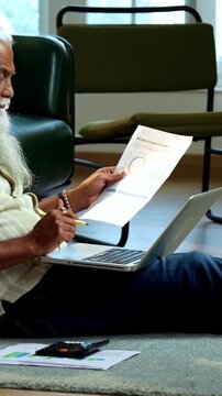 Indian mid age bearded man using laptop, calculator and reports for accounting sitting on carpet in modern home, stylish mature male managing finance, budget and financial statements indoors