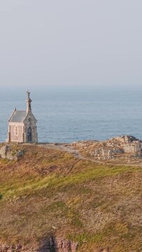 A small stone chapel stands alone on the rocky, grassy headland of Ilot Saint Michel, overlooking the calm sea along the Brittany coastline near Erquy, France.