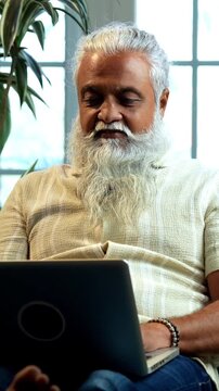 Indian mid age bearded man celebrating success sitting on sofa in modern home, stylish mature male expressing business profit achievement with raised fist and joyful expression indoors