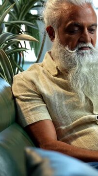 Indian mid age bearded man celebrating success sitting on sofa in modern home, stylish mature male expressing business profit achievement with raised fist and joyful expression indoors
