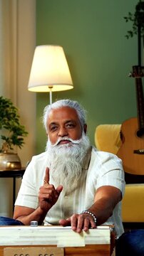 Indian mid age bearded man recording vocals music session on smartphone for online class at home, stylish mature tutor teaching Indian classical singing with harmonium in modern studio