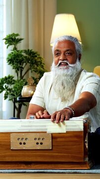 Indian mid age bearded man recording vocals music session on smartphone for online class at home, stylish mature tutor teaching Indian classical singing with harmonium in modern studio