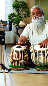 Indian mid age stylish bearded man taking online Tabla musical instrument playing class at home, experienced classical percussion tutor streaming lesson on smartphone in modern studio
