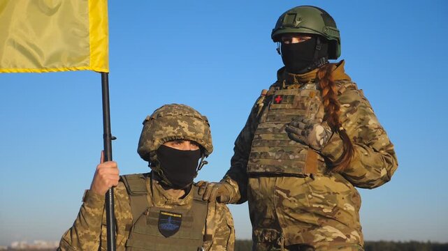 Portrait of young soldiers in camouflage uniform with lifted blue-yellow banner at countryside. Military couple of ukrainian army with a waving flag of Ukraine posing on camera against blue sky