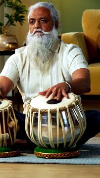 Indian mid age stylish bearded man practicing playing Tabla musical instrument at home, dedicated classical percussion performer doing daily riyaz session in modern home studio interior