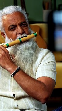 Indian mid age stylish bearded man teaching playing wooden flute musical instrument at home using smartphone, experienced classical bansuri tutor conducting online class in modern studio