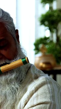 Indian mid age stylish bearded man practicing playing wooden flute musical instrument at home, dedicated classical bansuri performer doing daily riyaz session in modern home studio interior