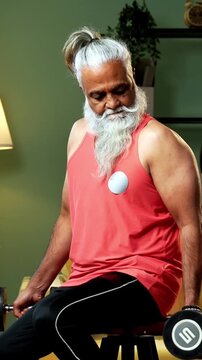 Indian mid age stylish bearded man doing exercise using dumbbell at home, fitness conscious mature male performing weight training workout showing strength, agility determination in modern interior