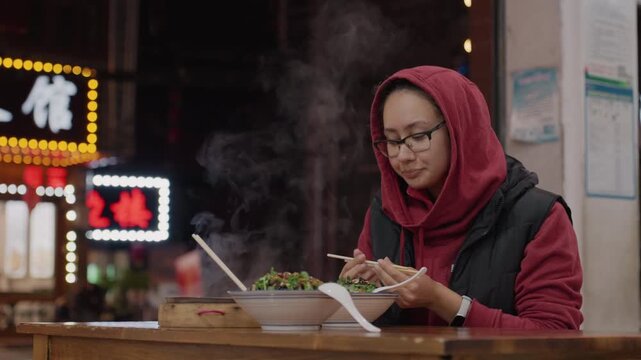 Hooded asian woman eating noodles under neon at night market steam rising from bowl, chopsticks and wooden table, thoughtful expression, candid street food scene, student pausing from night walk