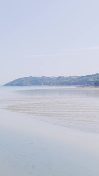 A serene summer scene of Binic beach on the Brittany coast in France, showing a wide sandy shoreline at low tide with gentle ripples in the wet sand and calm sea.