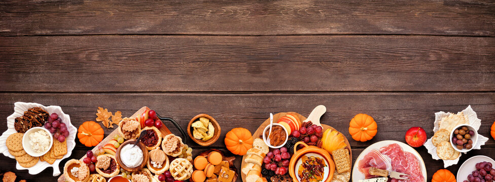 Autumn or Thanksgiving appetizers, sweets and grazing boards. Top down view bottom border on a dark wood banner background. Pumpkin and apple snacks, charcuterie, pies, desserts.