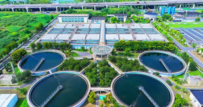 Drone view of a wastewater treatment facility with circular clarifier tanks, rectangular processing basins, surrounding greenery, and highway infrastructure in daylight.