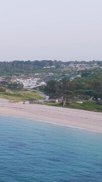 Aerial view of Plage Saint Michel in Erquy, Brittany, France, showing turquoise sea, sandy and pebbly beach, coastal vegetation, parked cars, and a small town in the background.