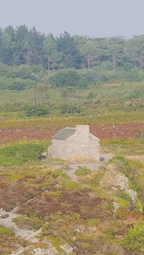 Aerial view of a historic stone guardhouse (corps de garde) standing alone on the wild heathland of Cap d'Erquy, surrounded by colorful summer vegetation and dense pine forest in Brittany, France.