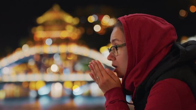 Night river scene female traveler warming hands near face visible breath in chilly air illuminated bridge lights create vibrant bokeh glasses and headscarf expressive gestures suggesting story