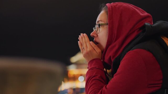 Night waterfront gaze female observer hopeful hands clasped near face looking up toward illuminated architecture soft reflections on water quiet meditative mood scarf and backpack hint travel