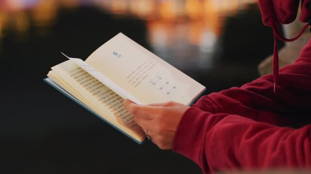 Night waterfront hands flipping book pages, red hoodie sleeve and closeup of fingers scanning text, soft reflections on water and neon bokeh, slow steady walking pace, focused nocturnal study