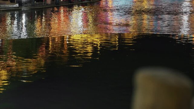 Night water reflections on river edge, colorful neon lights ripple across dark surface, blurred bollard in foreground, wet pavement gleam, tranquil urban atmosphere ideal for night walking scenes
