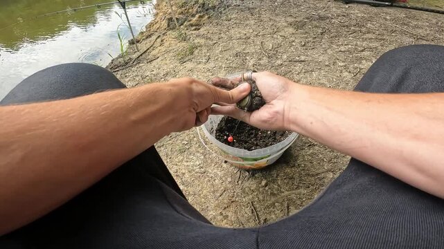 POV Angler hands mixing groundbait for feeder fishing in a plastic bucket next to a lake. Preparation of fishing bait for catching fish.