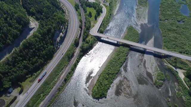 Top-down aerial view of the Matapedia River alongside a highway and railway, with a concrete beam bridge crossing the river corridor.