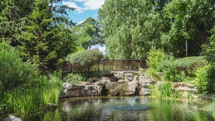 Fototapete Badezimmer Lush green park landscape with a small fountain pond, rocky waterfall feature, and wooden bridge under a sunny blue sky  © Вита Курило