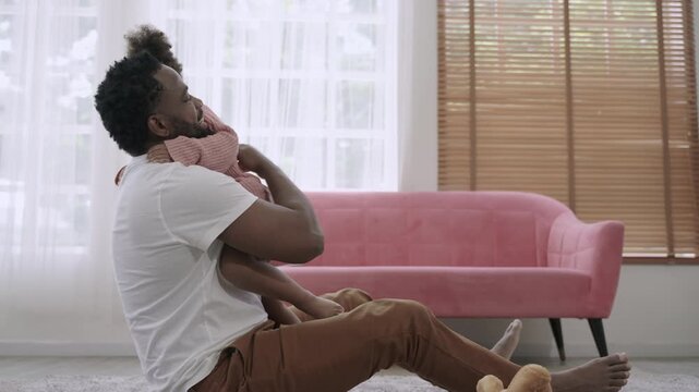 A cute African American child daughter is running and smiling to embrace her father while sitting and waiting on the carpet floor, happy and bonding in the living room at home. Father's Day concept. 
