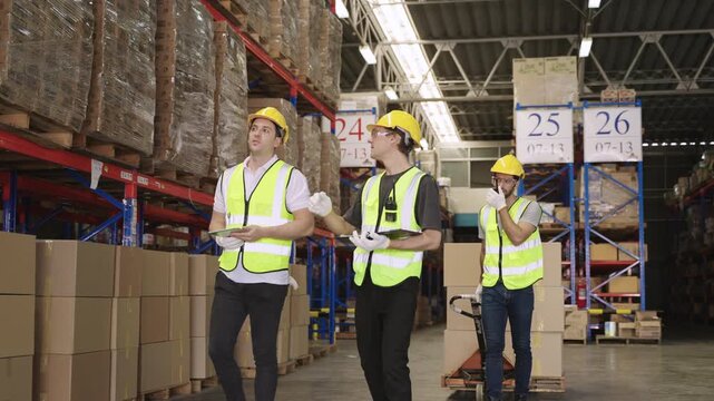 Teamwork of men warehouse workers in yellow helmets holding a tablet, working, talking, discussing, planning, inventory, moving cardboard boxes by hand pallet truck in logistics distribution factory. 