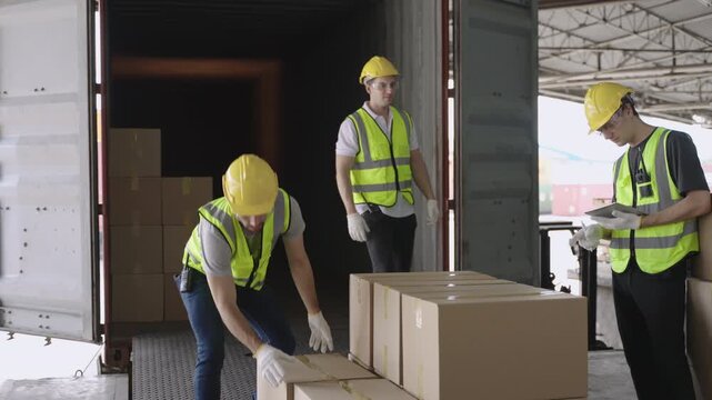 Team of young men warehouse workers in uniform wearing yellow safety helmets helping, holding, and carrying cargo boxes for loading into a container before delivery in a logistics distribution factory