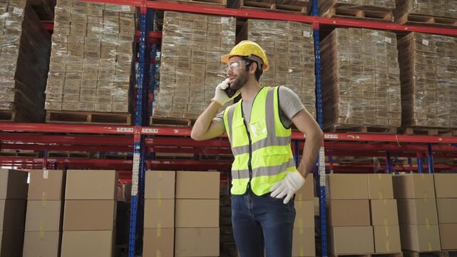 Young man warehouse worker in uniform wearing a yellow safety helmet and goggles, standing, using a smartphone, talking, discussing while checking inventory stock in a logistics distribution factory.