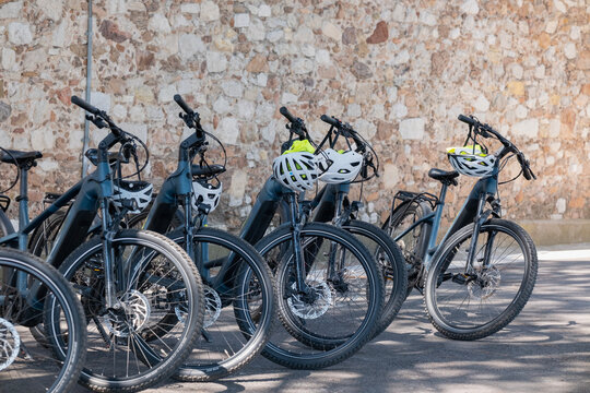 Several electric bicycles with helmets are parked in a row against a weathered stone wall, ready for riders exploring the surrounding area