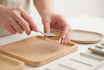 Obraz premium Hands using a small palette knife to fill a plastic watercolor pan with brown paint over a wooden tray