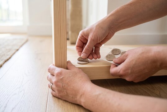 Hands peeling a round felt protective pad from a sheet and applying it to the bottom of a wooden furniture leg