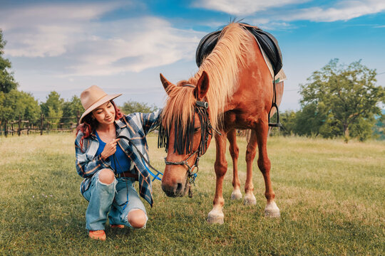 Tender moment as a smiling young woman kneels beside her saddled chestnut horse on a sunlit green pasture. Equestrian lifestyle concept.