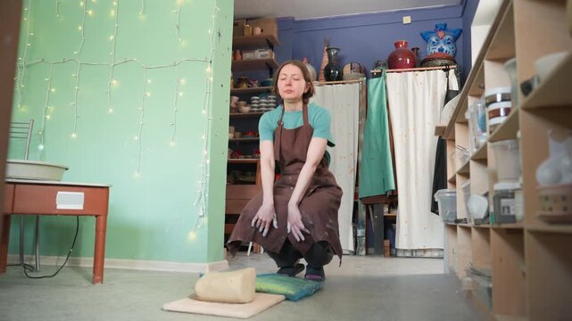 White woman preparing clay in studio, standing beside shelves of ceramics, methodical kneading and wedging on floor pad, brown apron splattered with clay, jars of glaze and tools on table, soft