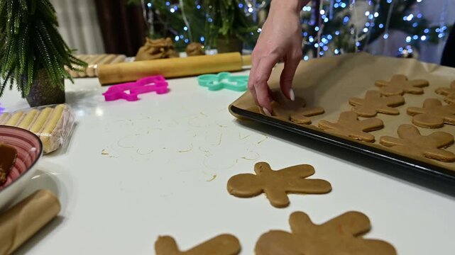 Beautiful hands carefully place gingerbread men on a baking sheet. The cozy setting includes a Christmas tree and colorful garlands, capturing the festive spirit of the season.