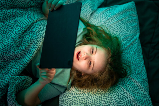 Child using tablet in bed. Kid holding tablet while lying at night. Teenager watching tablet screen in bed. Child playing games on tablet at night.