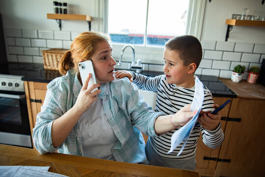 Stressed mother managing bills on phone with son in home kitchen