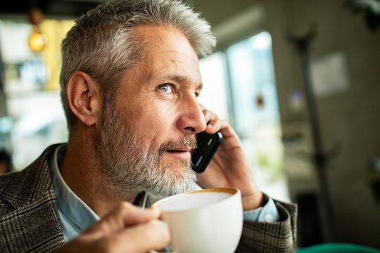 Mature businessman talking on smartphone while drinking coffee at cafe